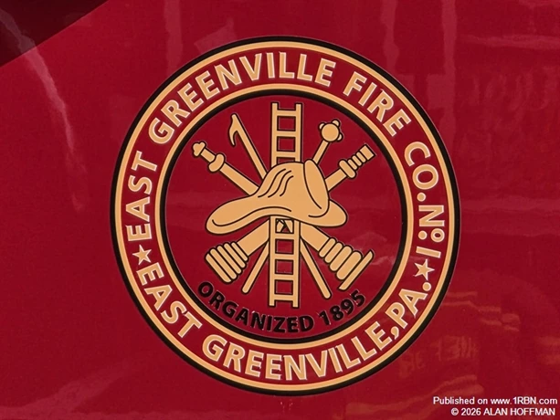 The emblem of East Greenville Fire Company No. 1 in Montgomery County, Pennsylvania, features traditional firefighting equipment, including a helmet, ladder and crossed tools, displayed in gold against a red background. The insignia prominently displays the company