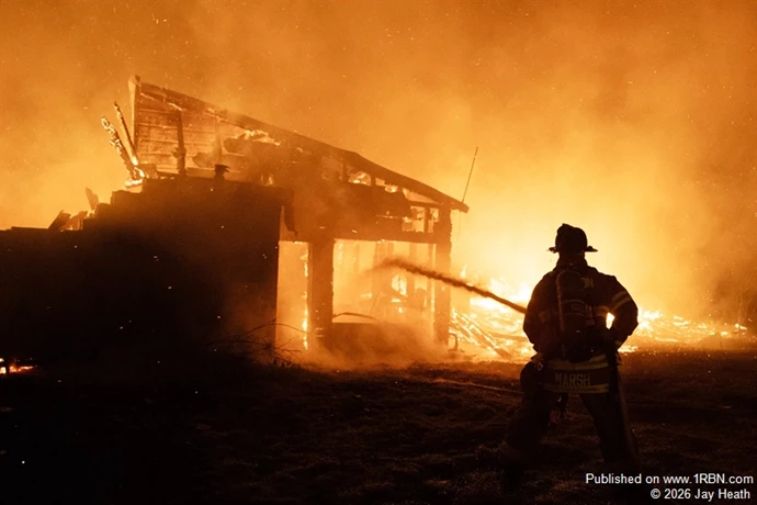 A firefighter operates a handline as flames engulf a historic barn during a second-alarm fire in Loudon, New Hampshire, on October 27, 2026. The approximately 200-year-old barn was destroyed, but firefighters from Loudon and surrounding communities prevented the fire from spreading to a nearby residence.